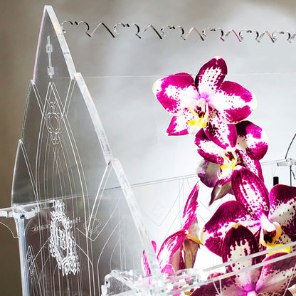Close-up view of intricate cutwork and engraving details on a Gothic-inspired terrarium, with a large blooming orchid in vibrant color.